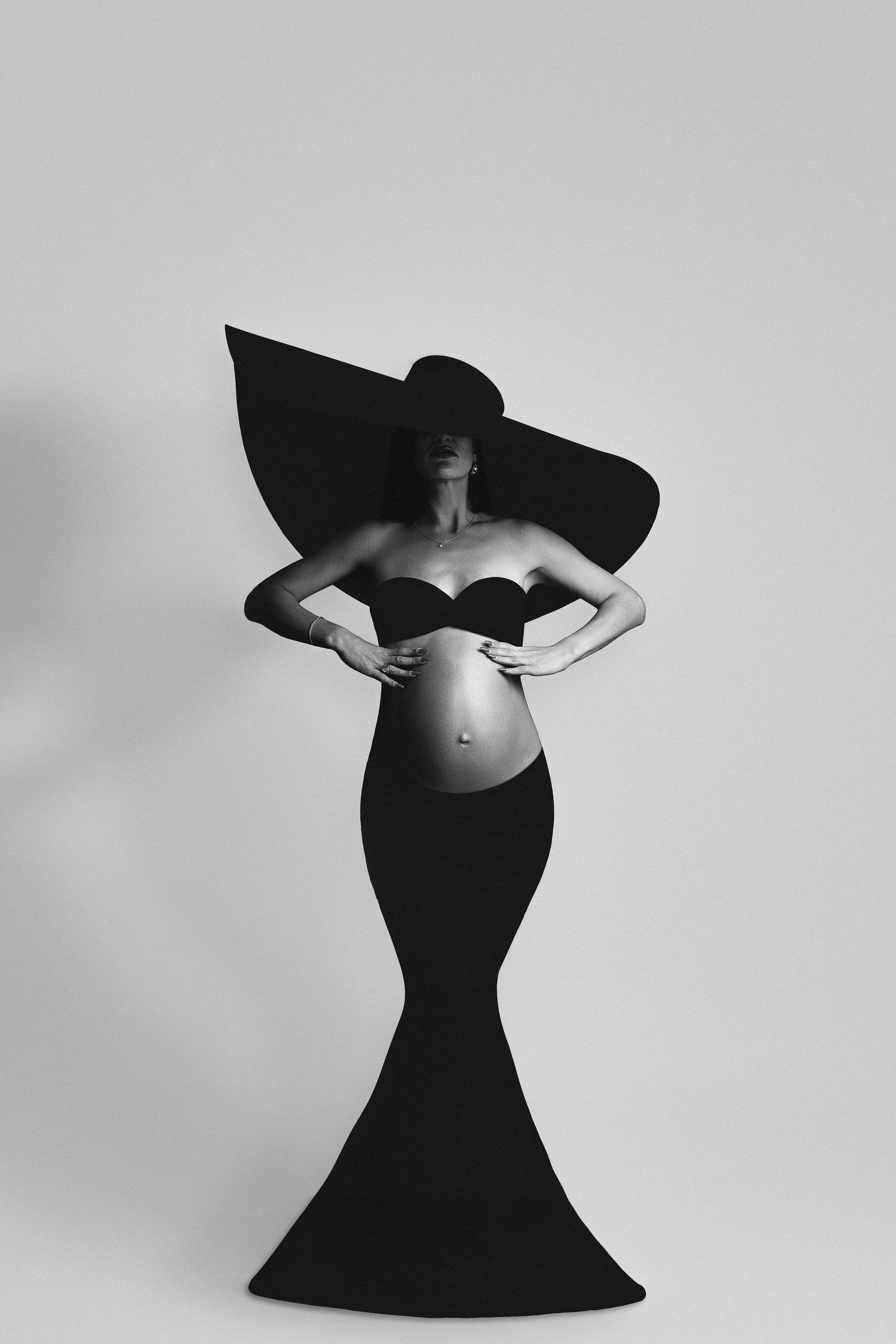Smiling pregnant woman in black dress cradling her baby bump, side profile studio portrait on gray background.