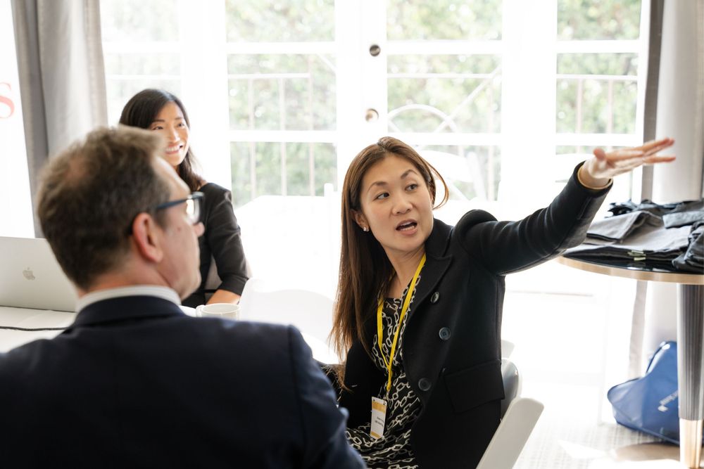A woman in a black blazer with a yellow lanyard gestures while speaking to colleagues in a bright office meeting.