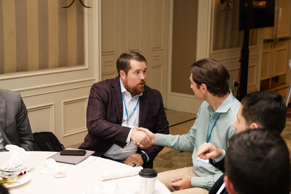 Two professionals shaking hands across a conference table at a business networking event.