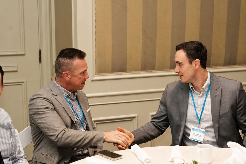 Two businessmen in suits shaking hands at a conference table during a professional networking event.