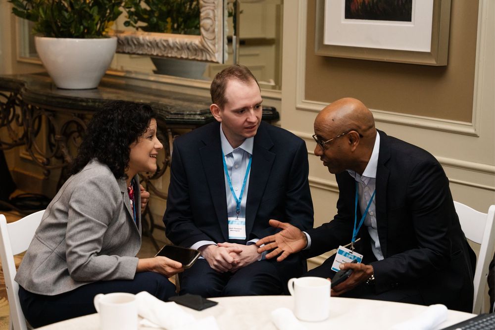 Three business professionals in formal attire engage in discussion at a conference table with coffee cups nearby.