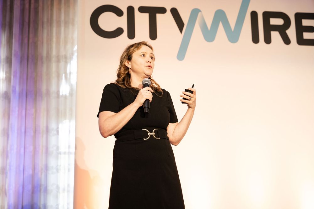Woman in black dress speaks on stage at Citywire event, holding microphone and phone.