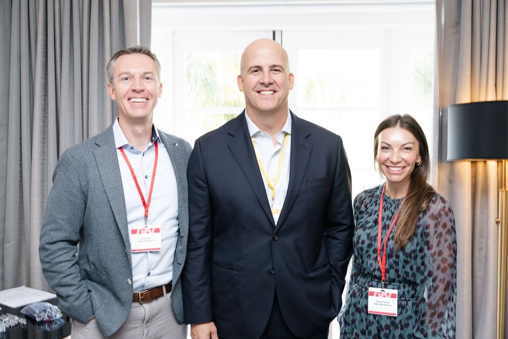 Three professionals wearing lanyards smile together at a corporate networking event in a modern venue.