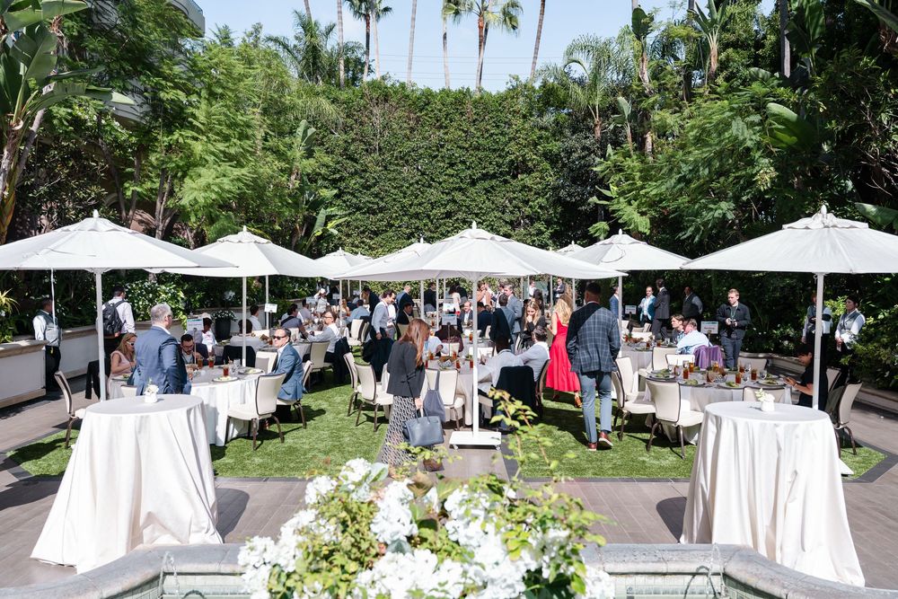 Elegant outdoor garden party with white umbrella tables, guests mingling on manicured lawn surrounded by lush tropical greenery.