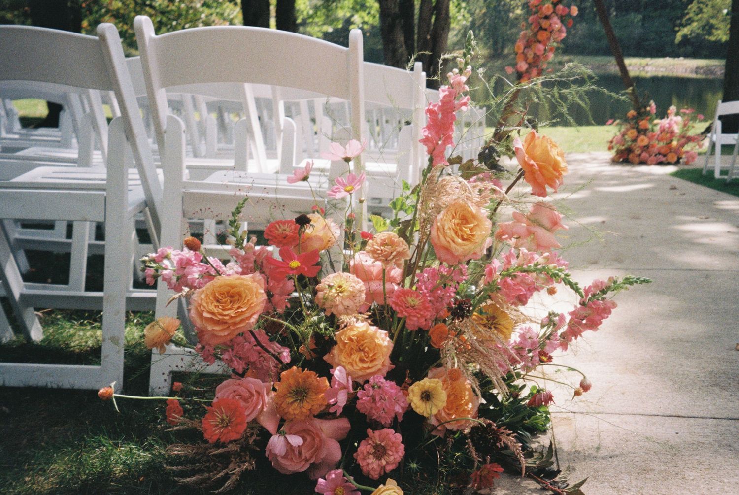Elegant outdoor wedding aisle lined with white folding chairs and lush pink and peach floral arrangements.