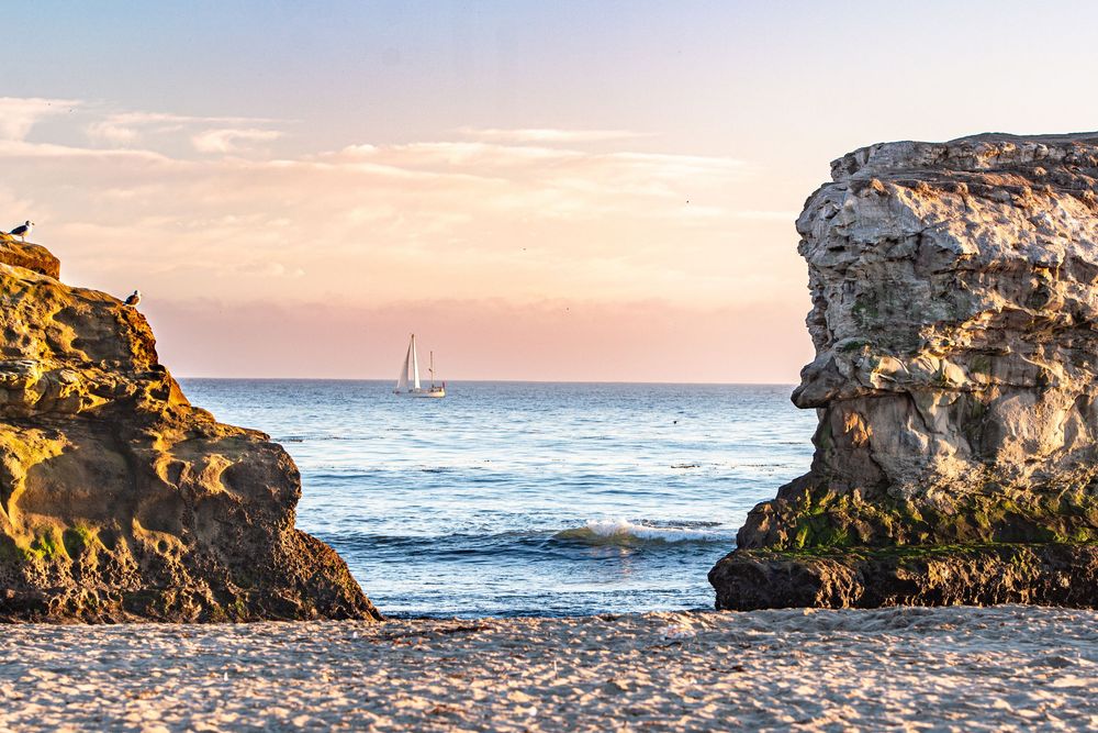 Scenic beach cove at sunset with rocky cliffs framing a sailboat on calm ocean waters.