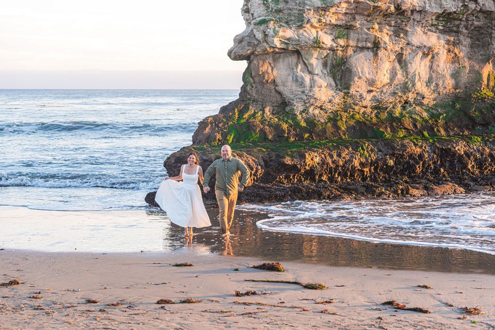 Bride and groom walking hand in hand on a sandy beach at sunset with rocky cliffs in the background.