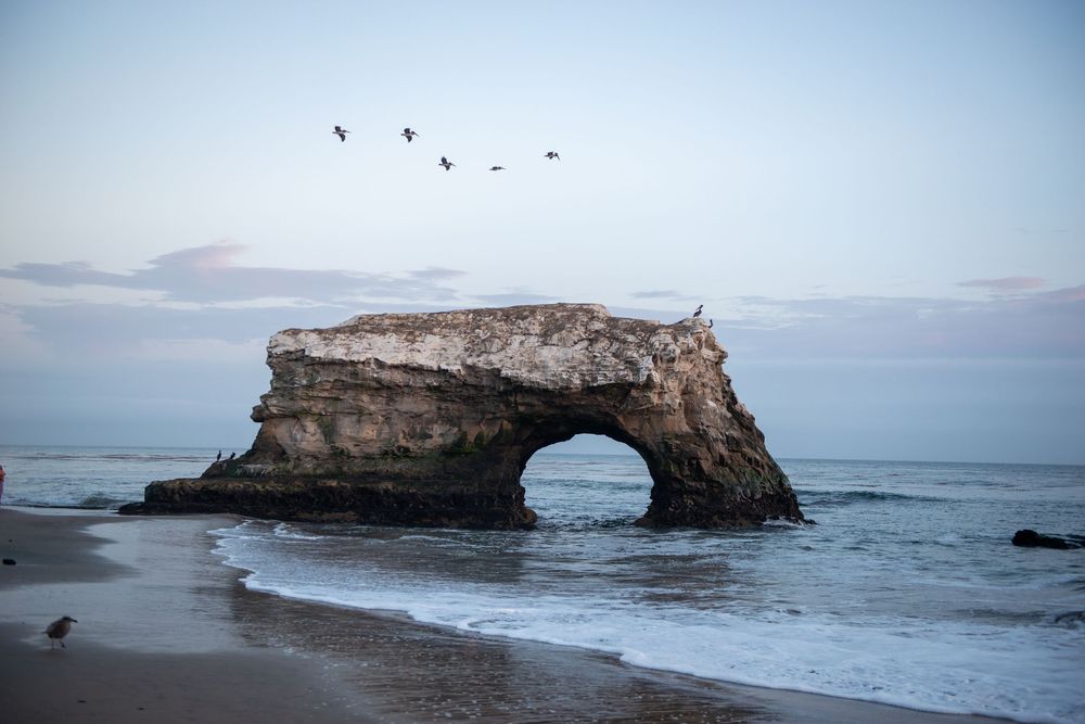 Natural rock arch formation rising from ocean waves on a sandy beach with birds flying overhead at dusk.