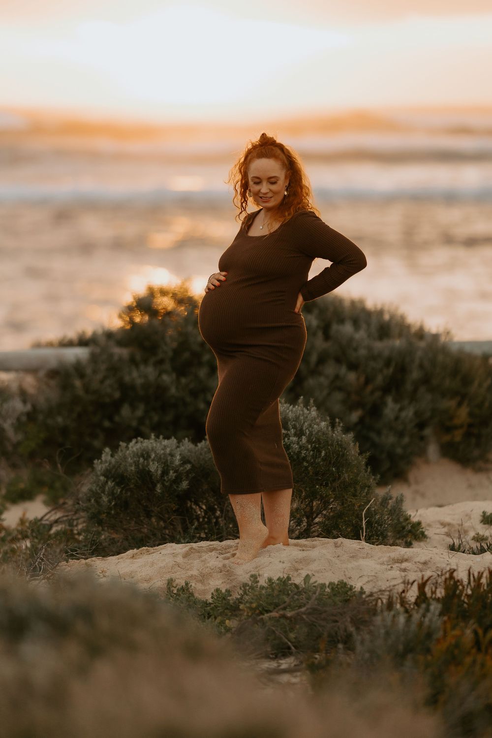 Pregnant woman in brown dress standing on sand dunes at golden hour sunset.