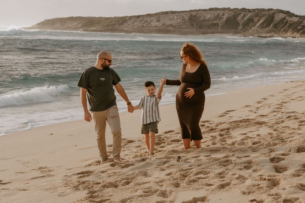 Pregnant woman and man hold hands with young boy walking on sandy beach with ocean waves in background.