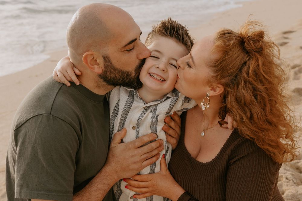Parents kissing their smiling young son on the cheeks at a sandy beach during golden hour.