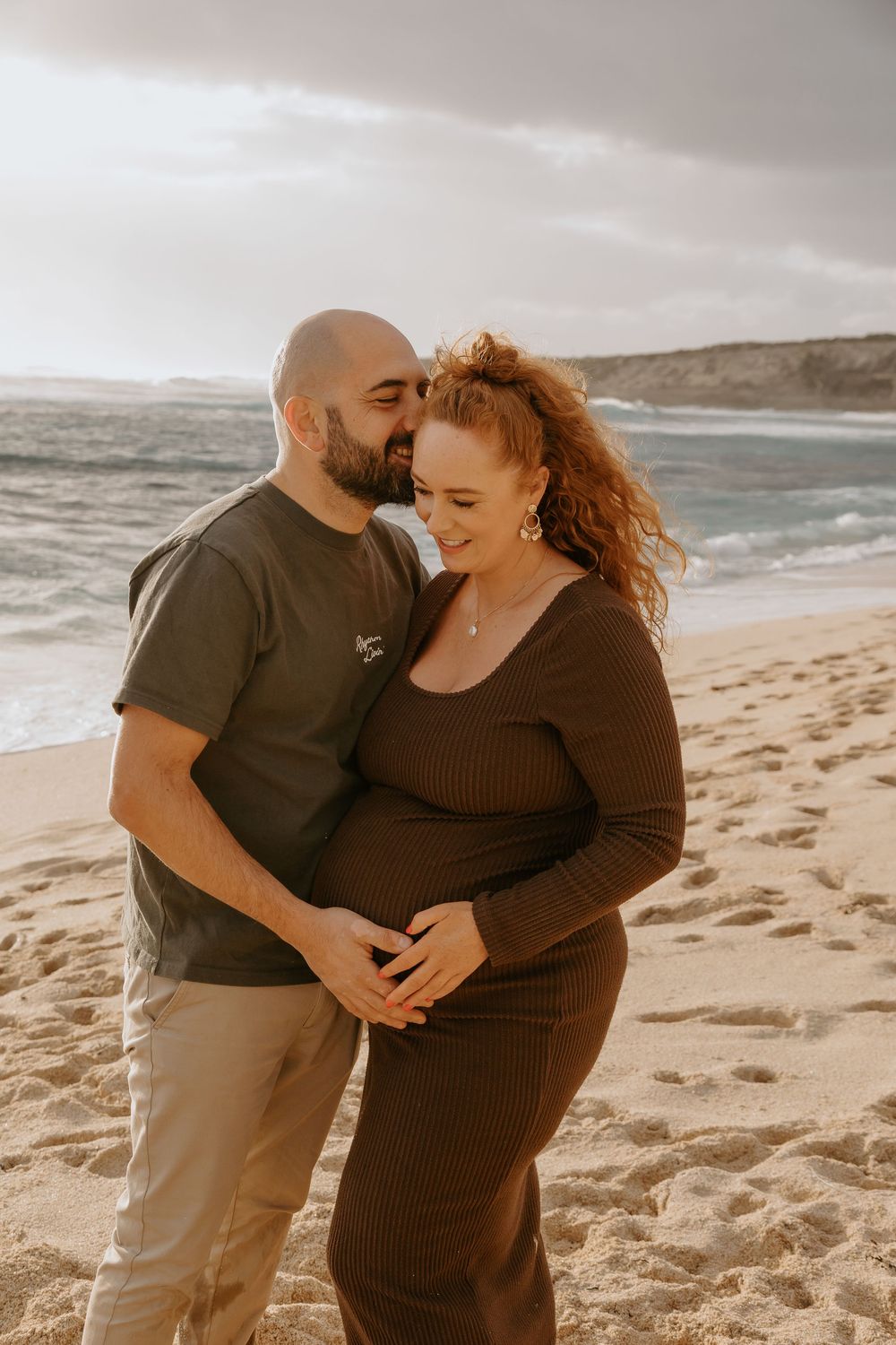 Couple shares tender beach moment, man embracing pregnant woman in brown dress at sunset.