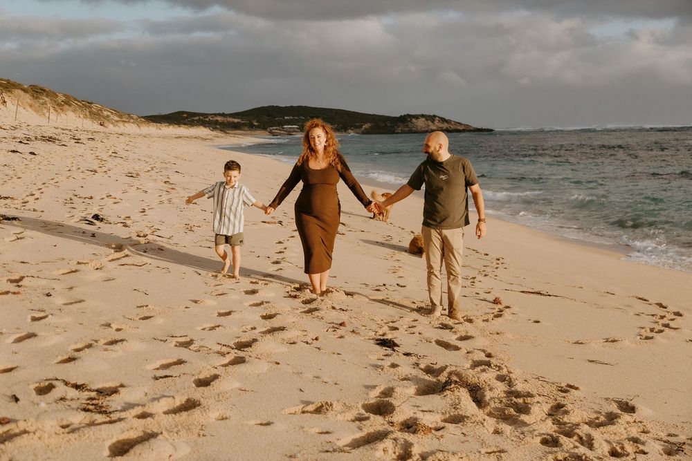 Family of three walking hand-in-hand on a sandy beach at golden hour, with ocean waves and dunes in the background.