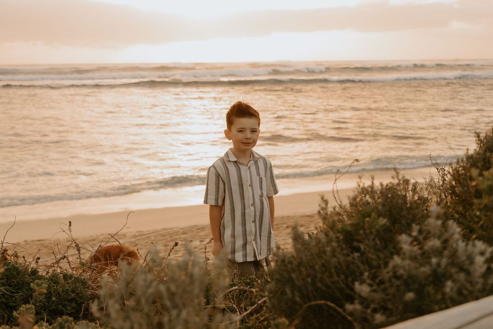 A young boy in a striped shirt stands on a sandy beach at golden sunset with ocean waves in the background.