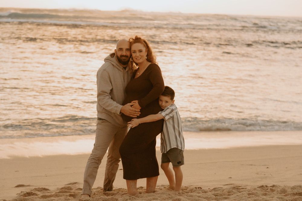 Family maternity photo on beach at golden hour, parents embracing pregnant mother while young boy hugs her side.