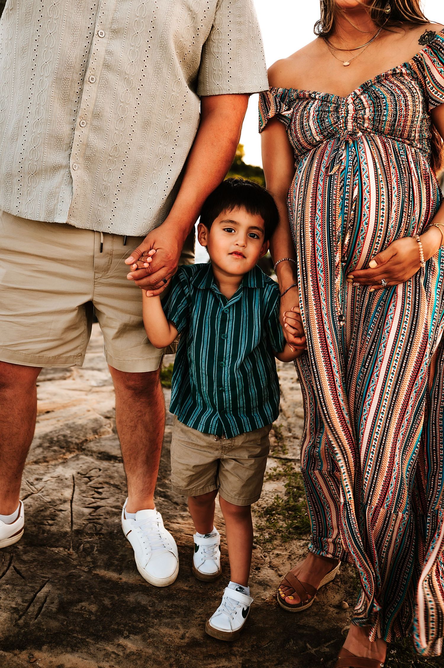 Close-up of pregnant woman in striped dress and young son holding fathers hands during family shoot.