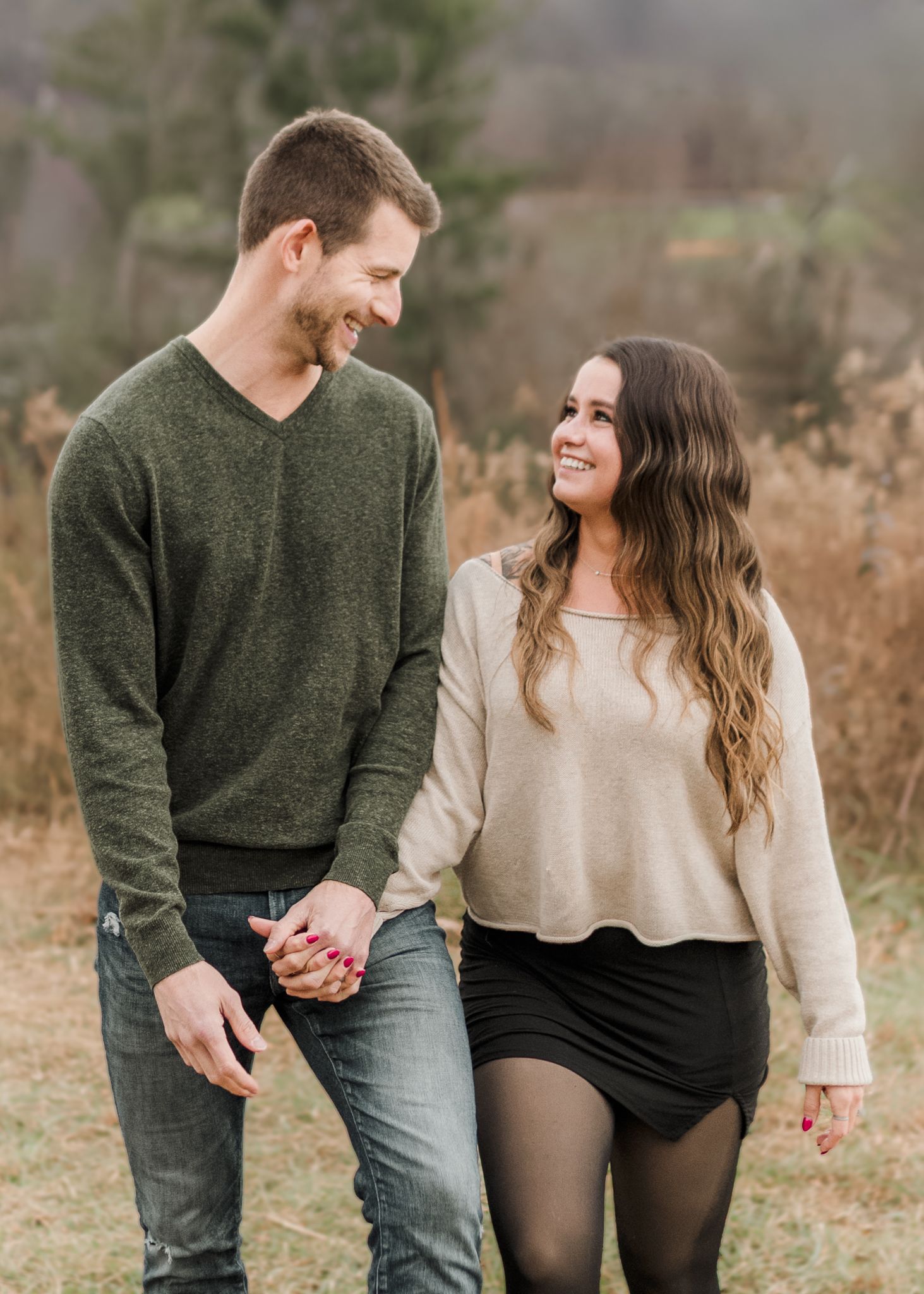 A smiling couple poses together in an autumn field, woman in cream sweater and man in green v-neck, with misty mountains behind.