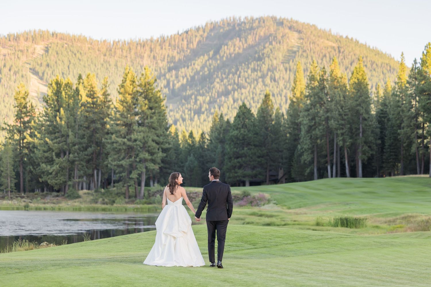 Bride and groom walk hand-in-hand on a lush green golf course with mountain and pine tree backdrop.