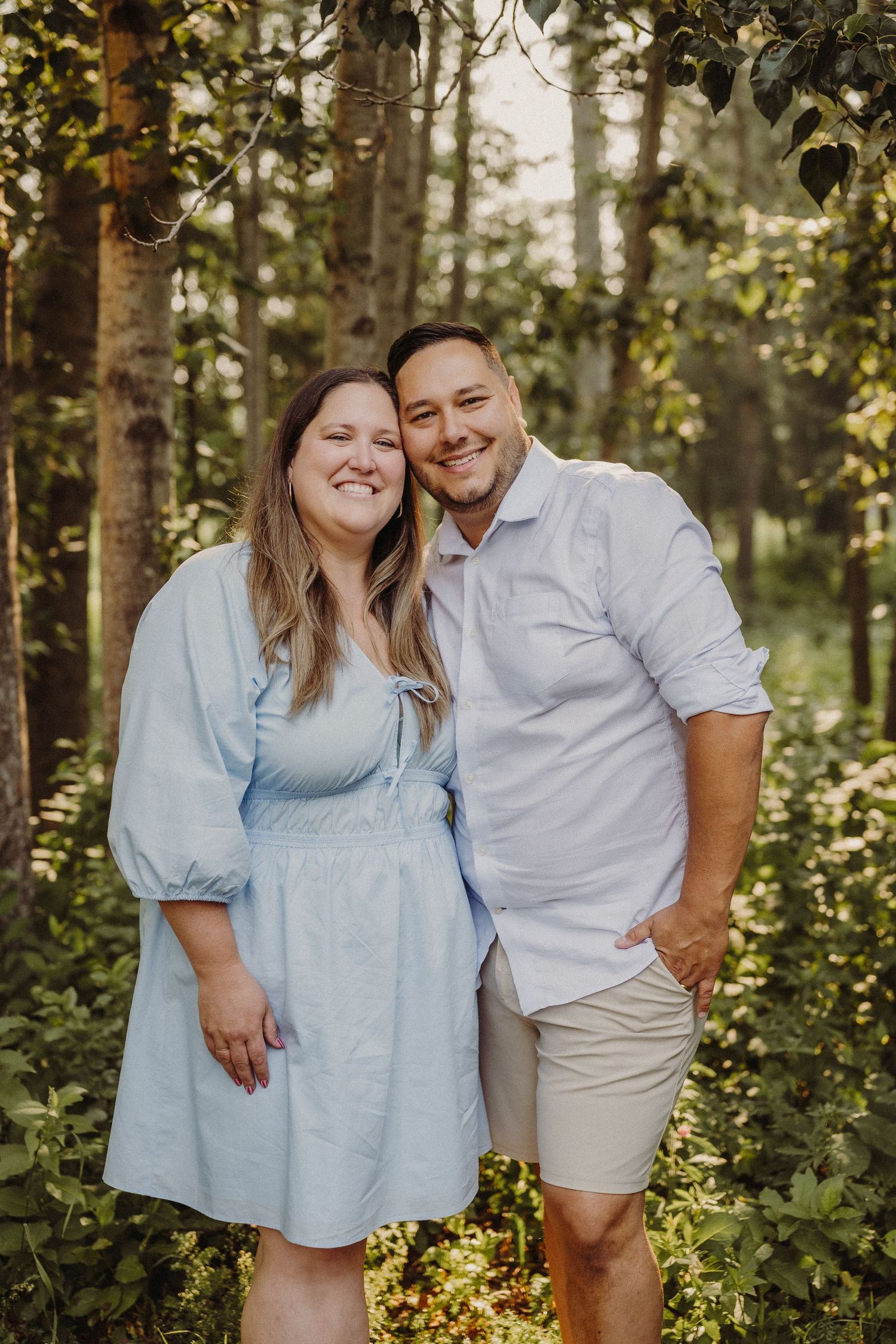 A smiling couple walks through shallow water, the man in a white shirt and the woman in a white slip dress, surrounded by greenery.