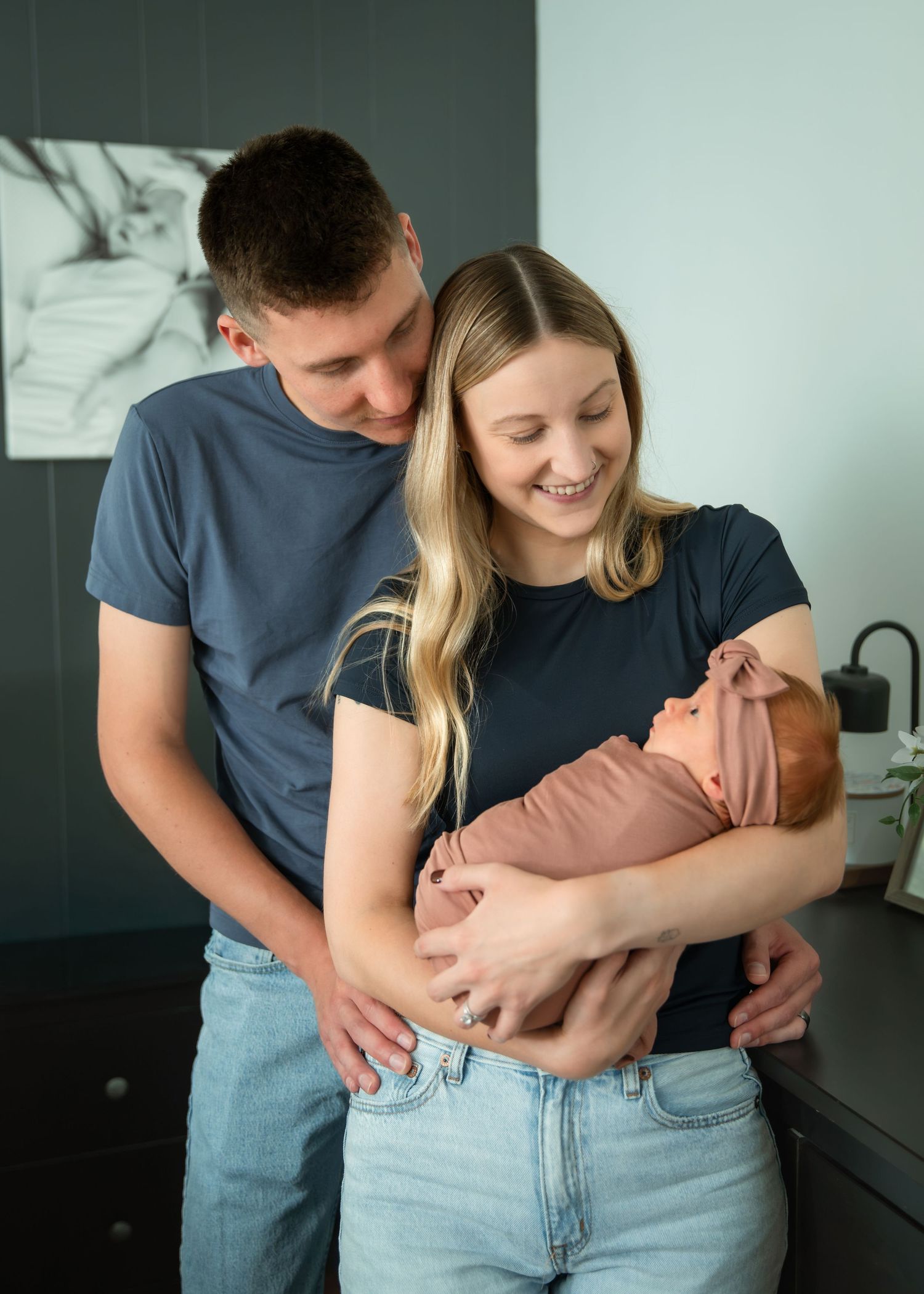 Young couple in matching navy shirts smiling at their newborn baby wrapped in a pink swaddle.