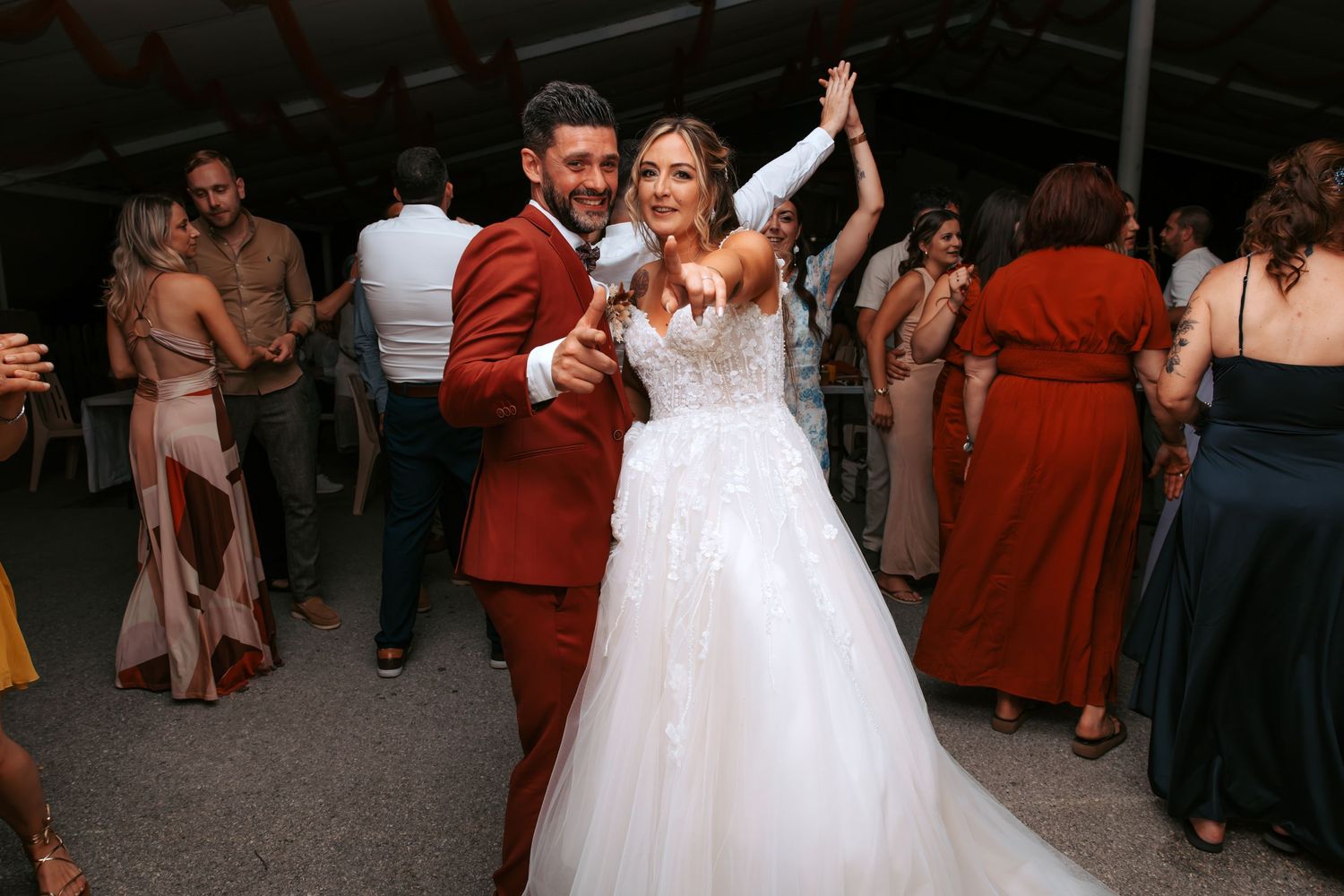 Bride in white lace gown and groom in red suit dancing joyfully at wedding reception with guests in background.