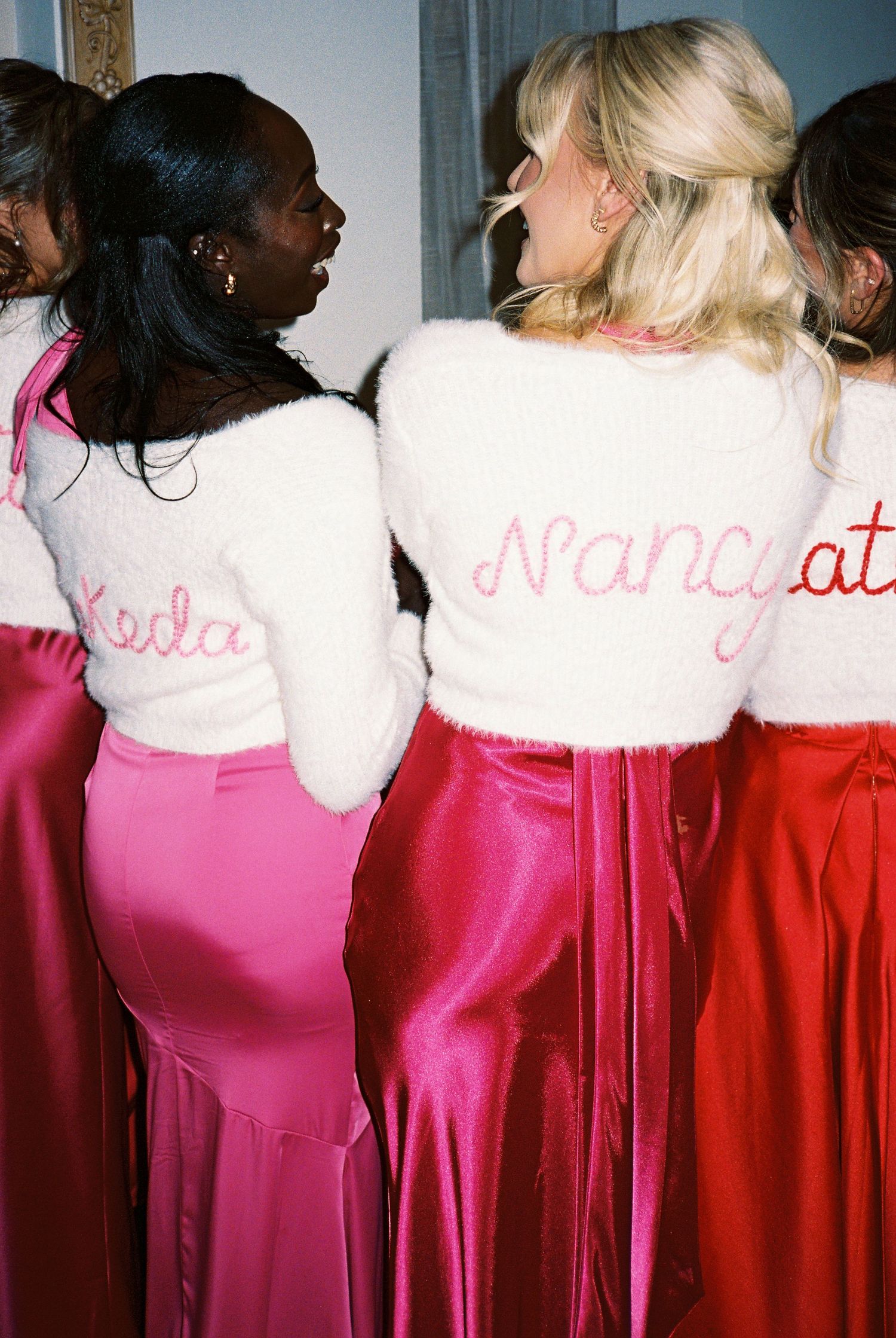 Bridesmaids in personalized white fuzzy sweaters with embroidered names and pink satin skirts at a wedding.