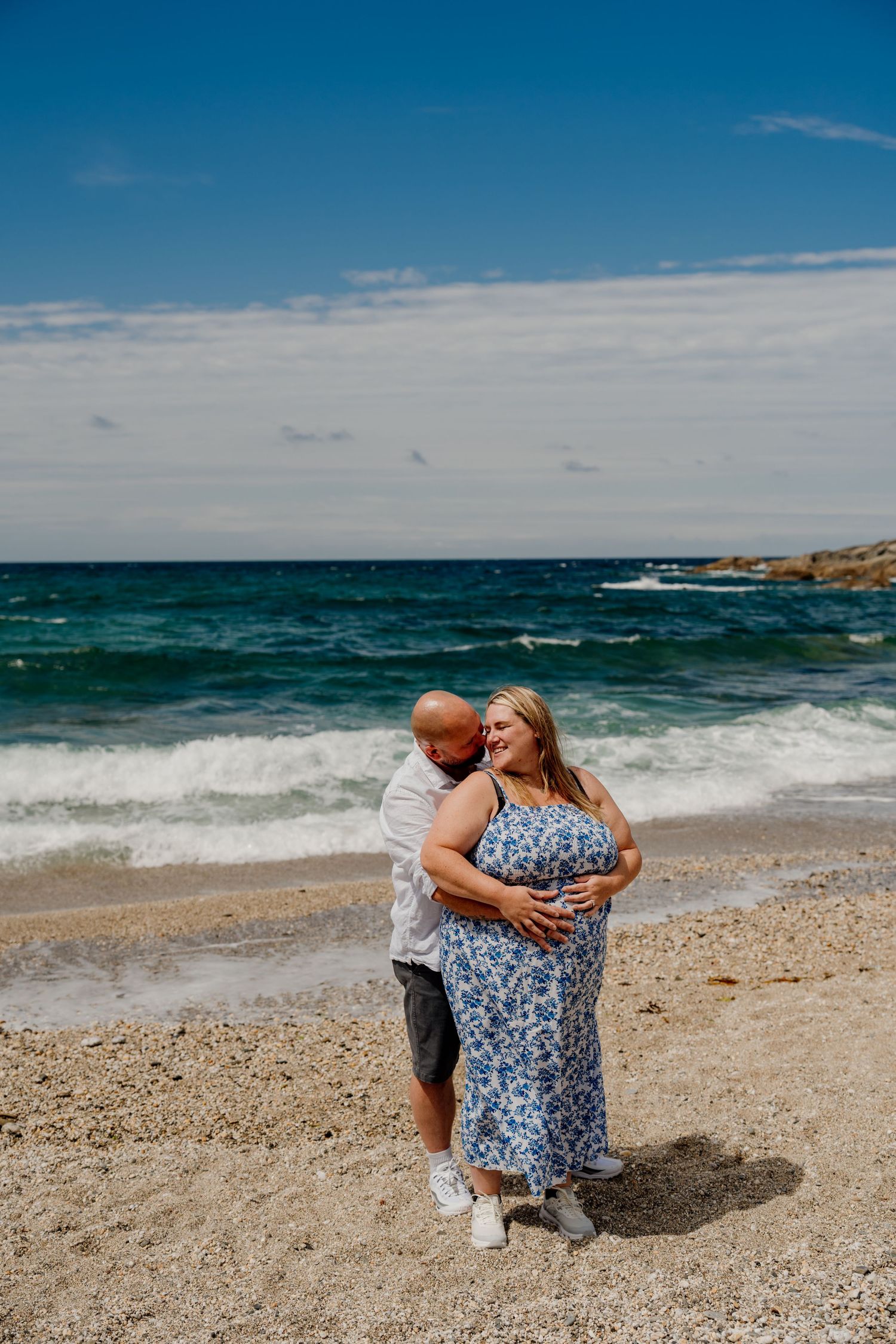 A couple embraces on a sandy beach, the woman in a blue floral dress cradling her baby bump on little fistral beach after proposal.