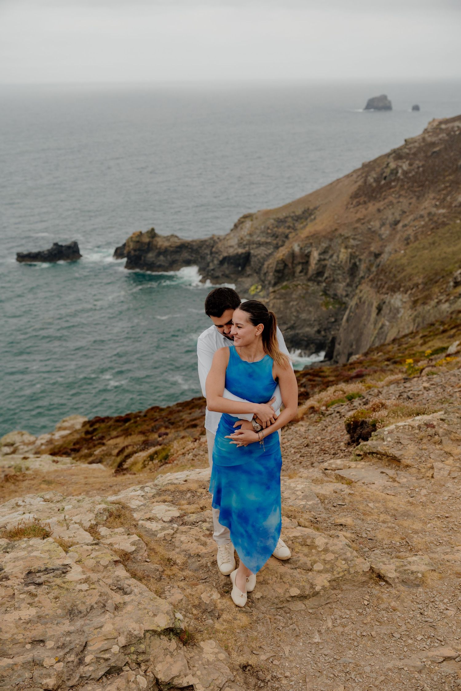 Couple embracing on st agnes head cliffs, woman in blue dress, ocean waves and misty sea stacks in background.