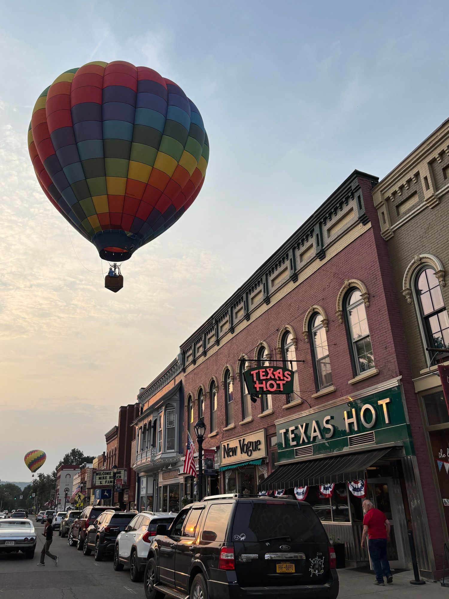 Colorful hot air balloon floating over Texas Hot restaurant on a brick-lined small town street at dusk.