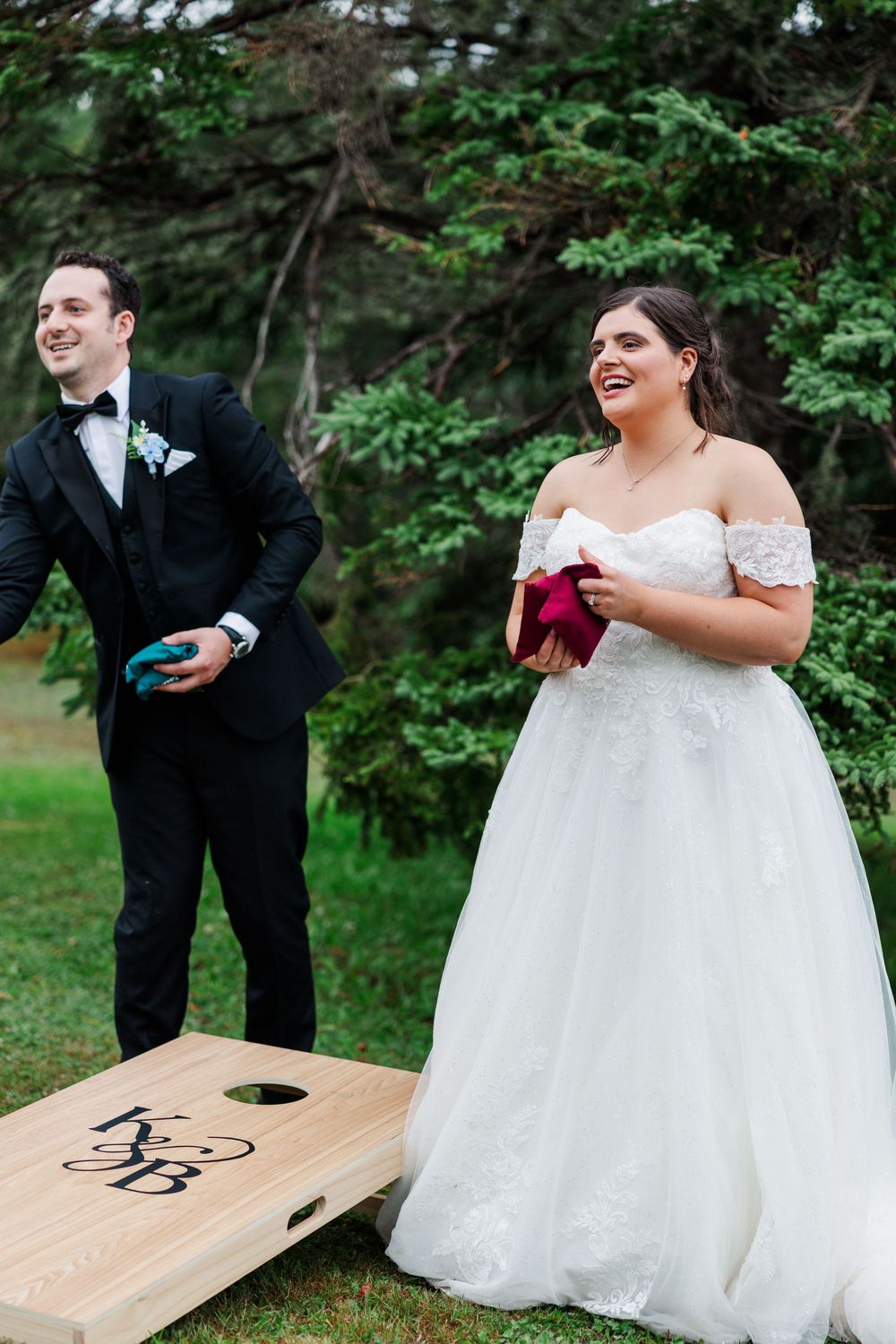 Bride and groom playing cornhole lawn game at outdoor wedding reception, bride in off-shoulder gown holding red bag.