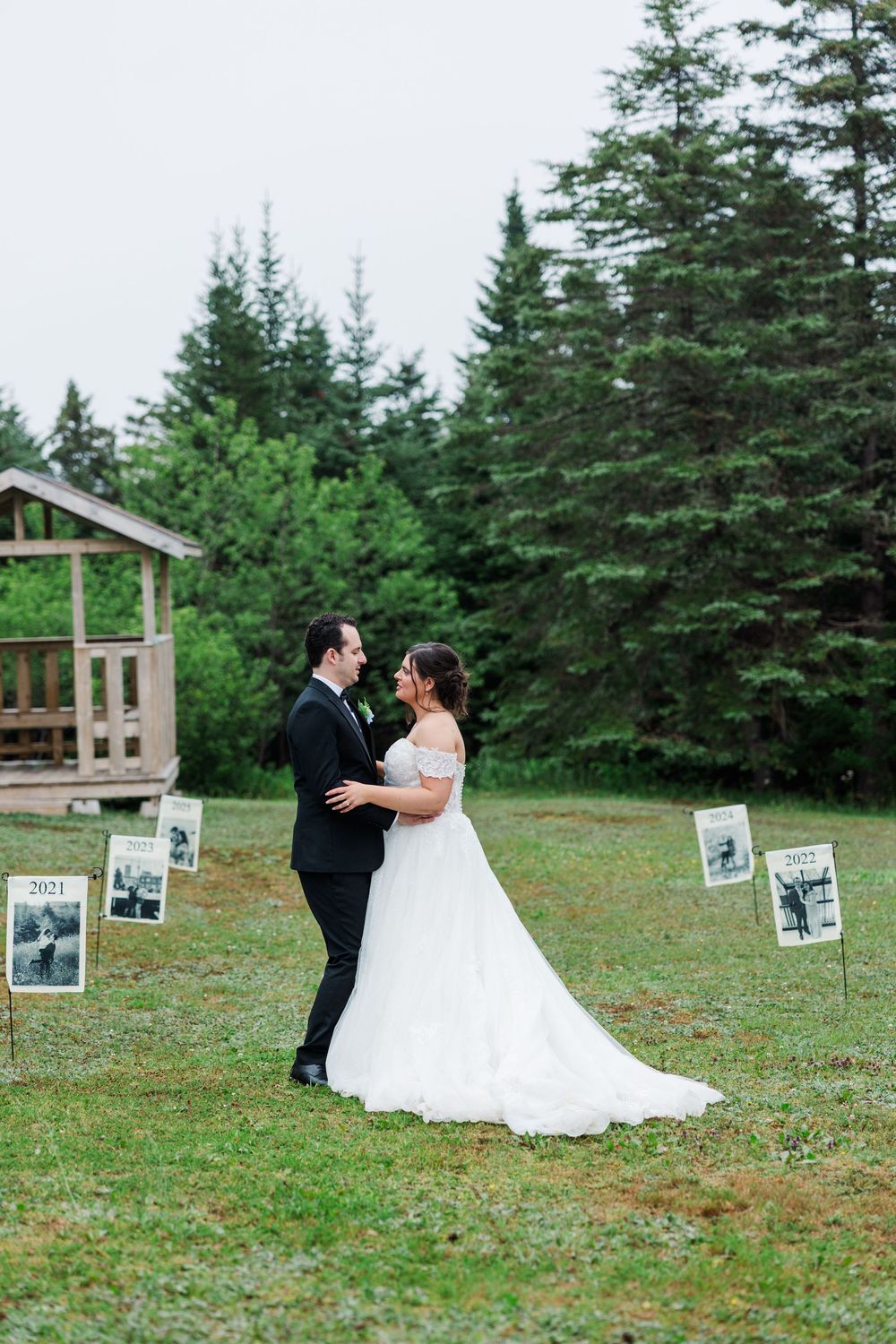 Bride and groom share a romantic moment outdoors surrounded by framed photos on grass with evergreen trees behind them.