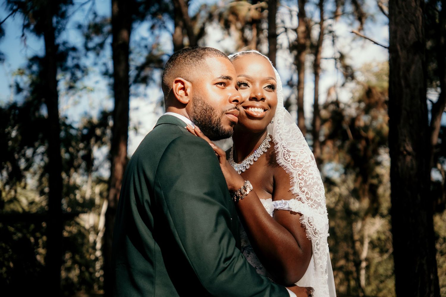A couple embraces in a forest setting, the groom in a green suit and the bride wearing a lace veil on their wedding day.