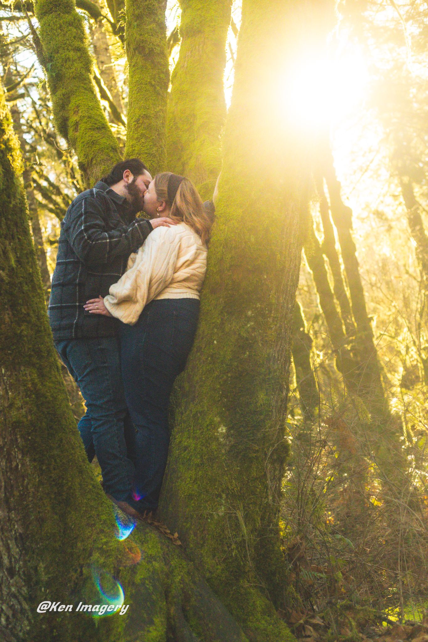 Couple sharing a romantic kiss against moss-covered trees in a sunlit forest, captured by Ken Imagery.