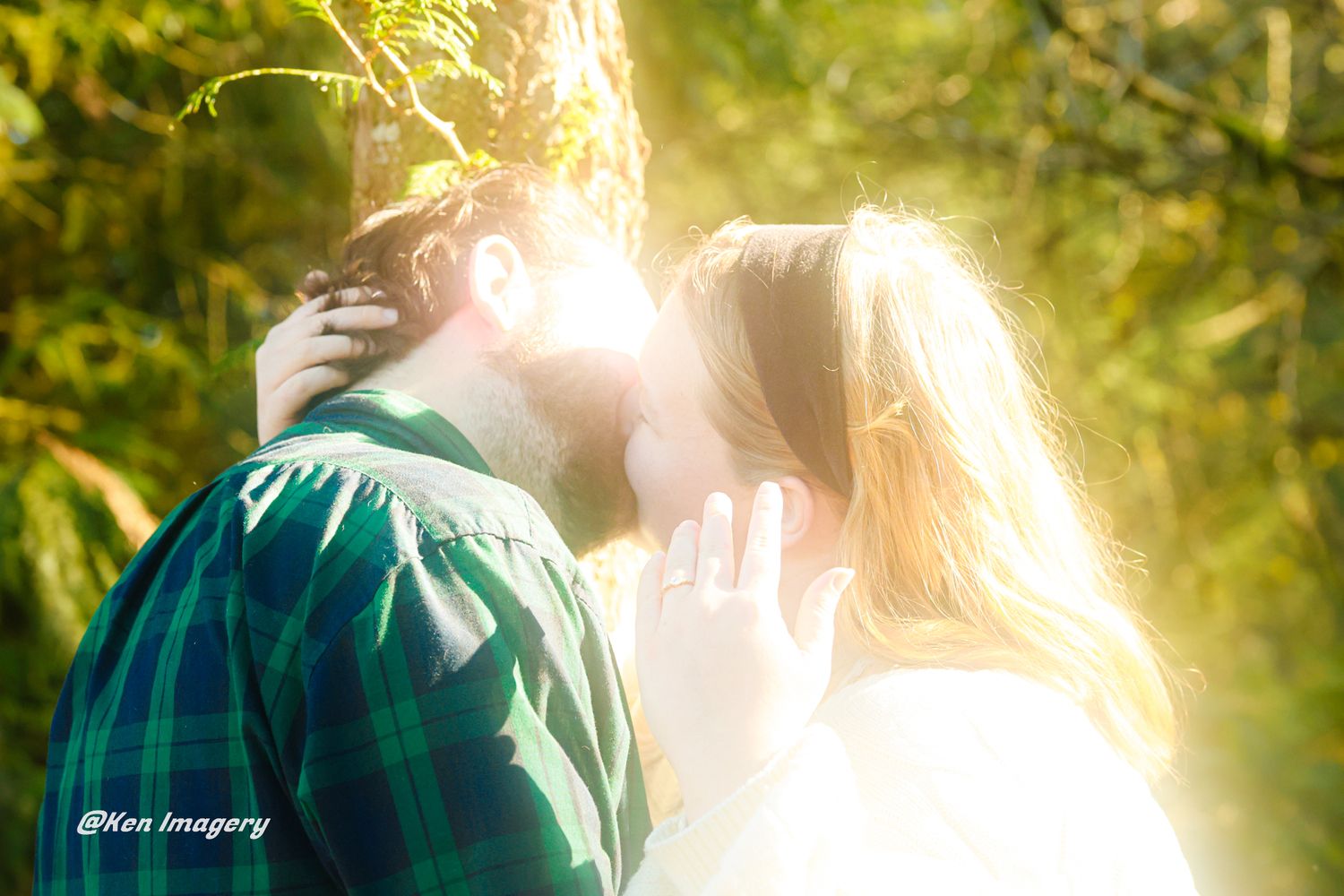 Couple sharing a romantic kiss near a sunlit tree in a lush green forest, captured by Hen Imagery.