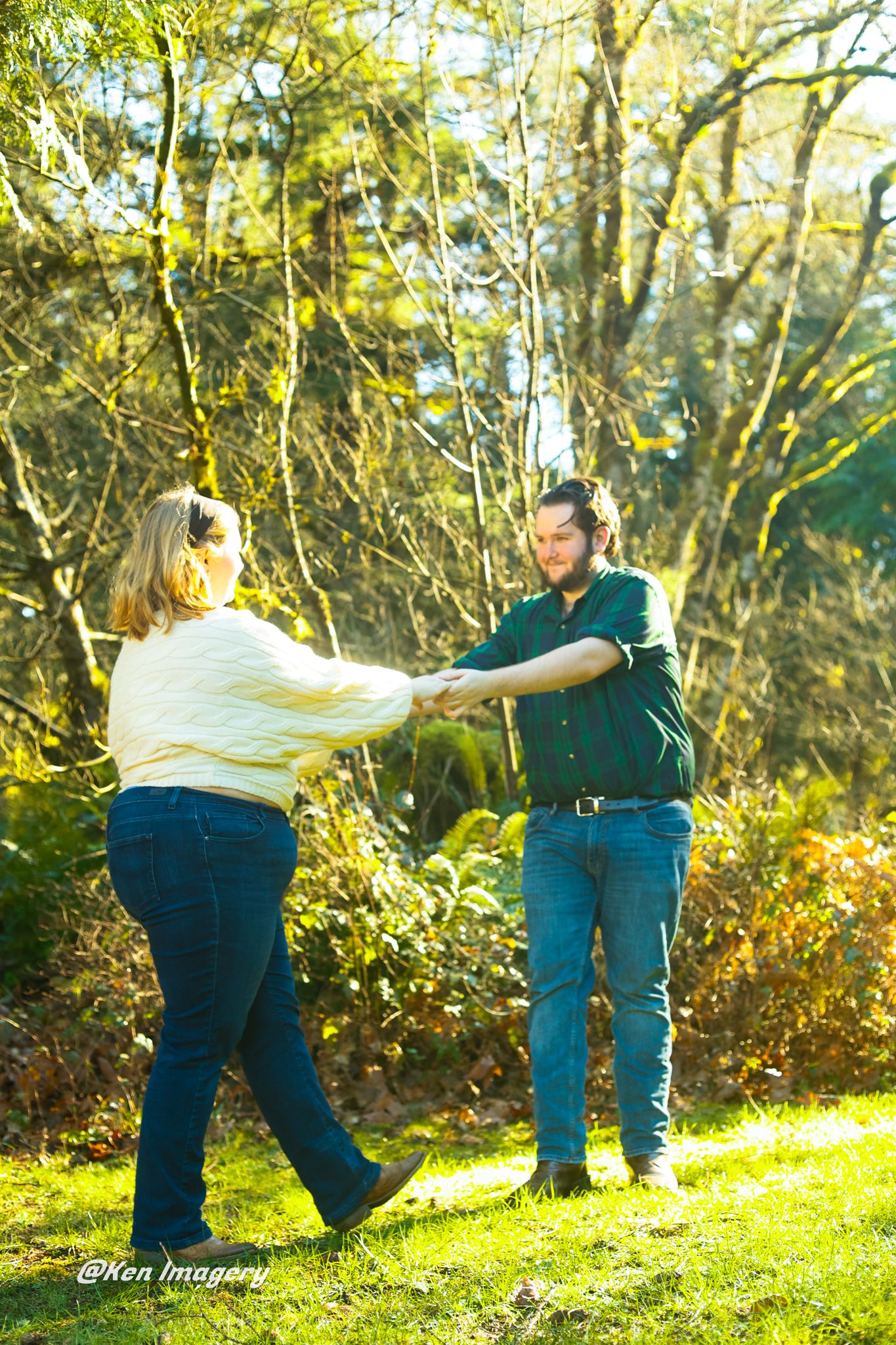 A couple holds hands and dances together on a sunny green lawn surrounded by trees in an outdoor garden setting.