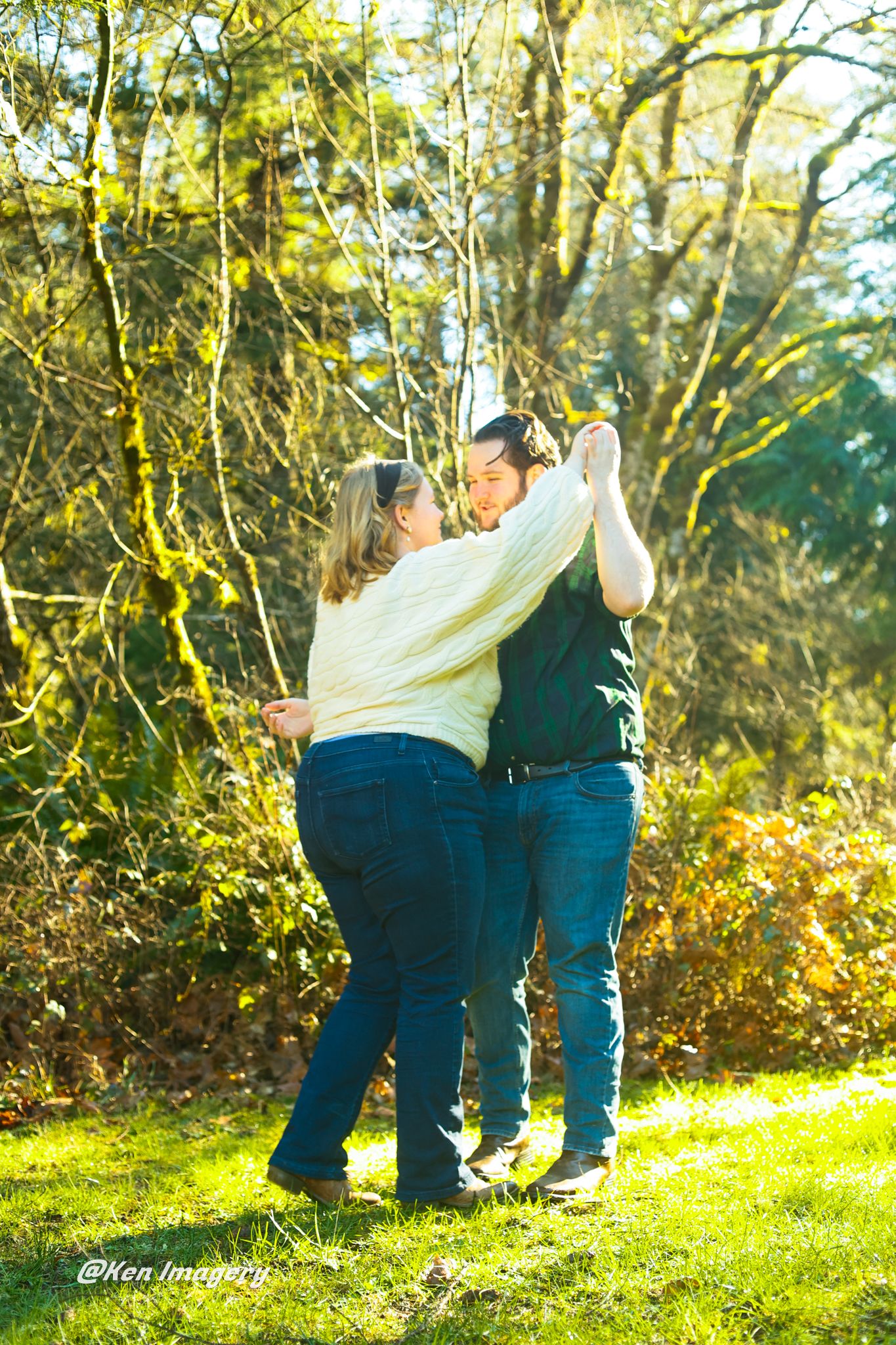 Couple dancing outdoors on green grass surrounded by sunlit trees in a romantic engagement photo session.