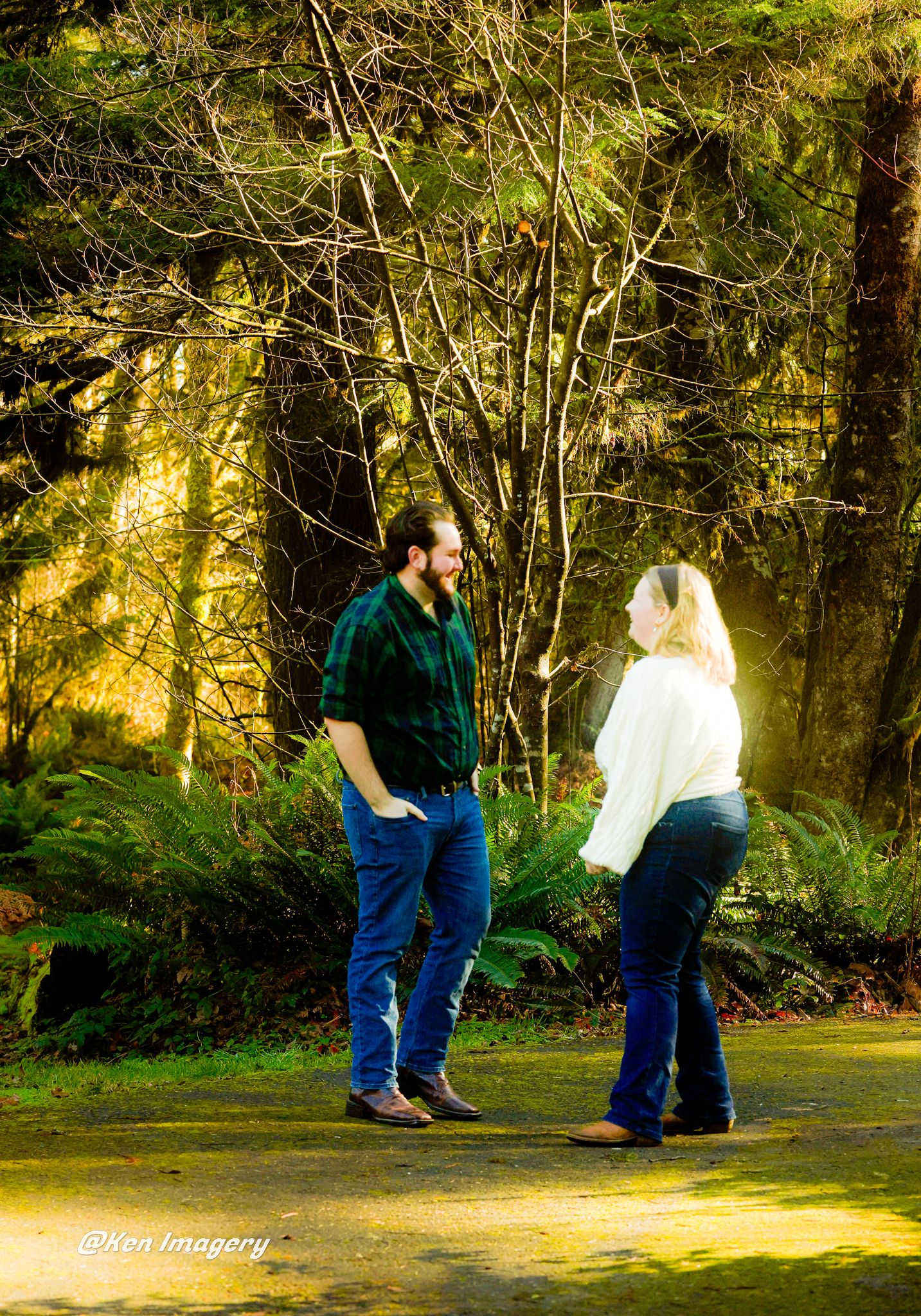 Couple sharing a romantic moment in a lush forest setting, man in green plaid shirt and woman in white top, both wearing jeans.