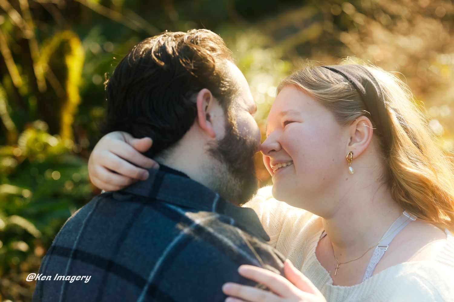 Couple sharing a romantic moment outdoors, woman smiling joyfully as they lean in close, surrounded by golden sunlit foliage.