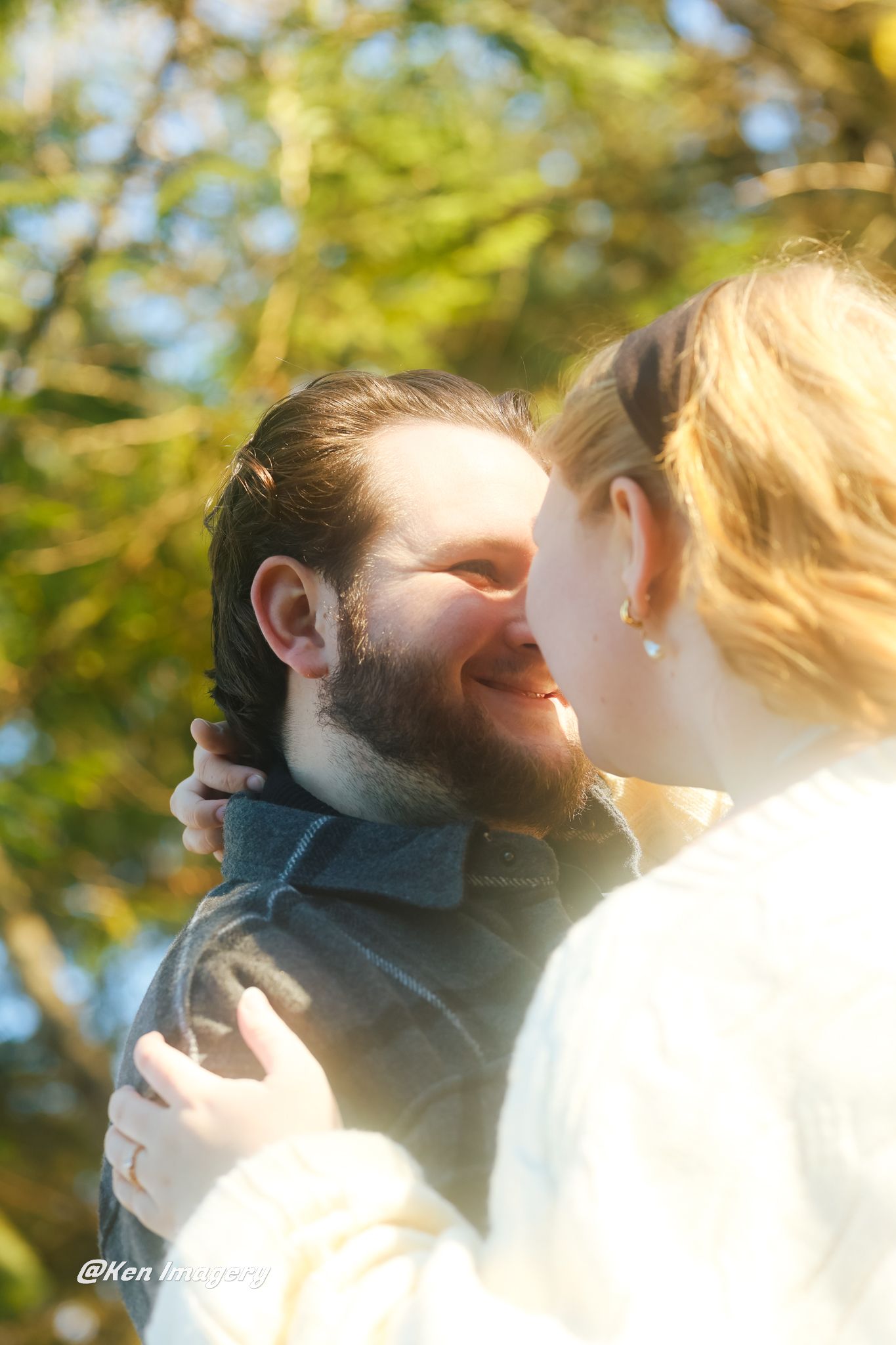 A bearded man smiles as a blonde woman embraces him outdoors with sunlit trees in the background.