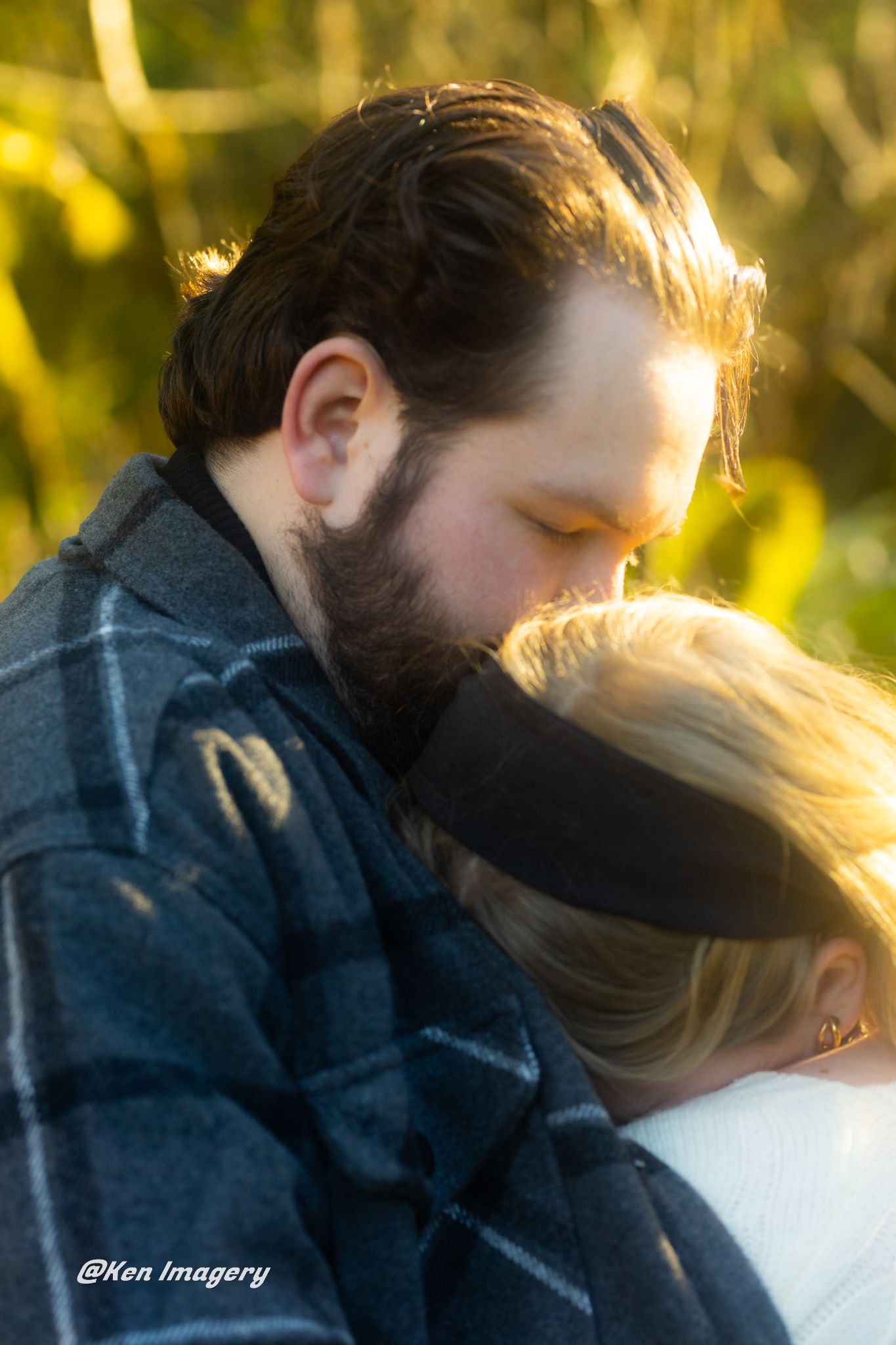 A bearded man in a plaid jacket tenderly kisses a blonde woman outdoors in warm golden light.