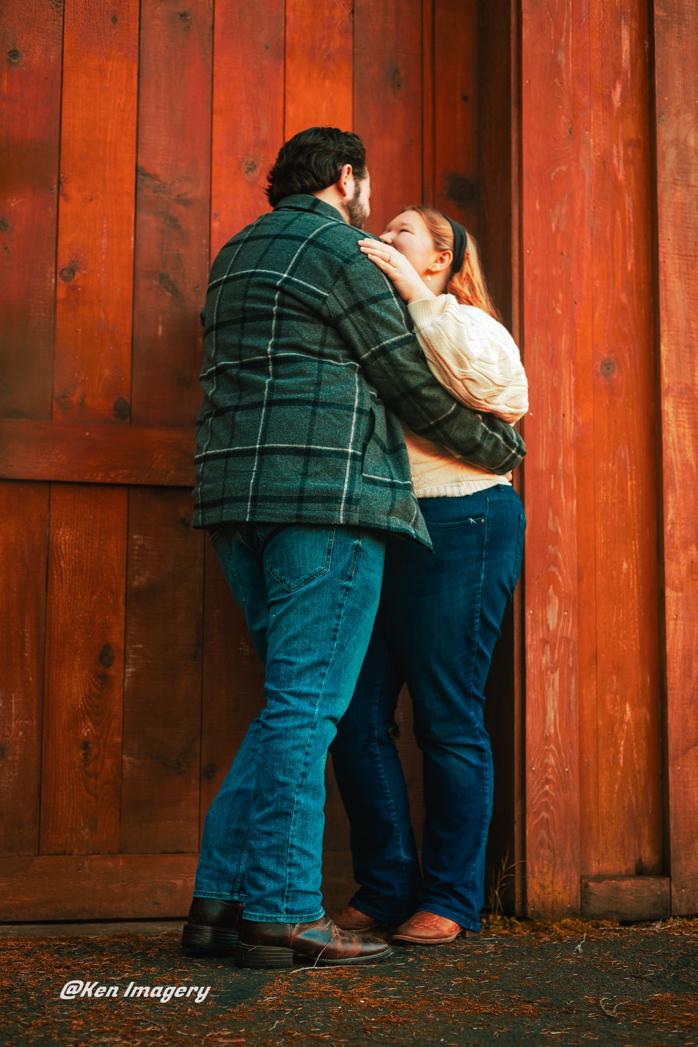 Couple embracing romantically against a rustic red barn wall, wearing jeans and plaid jacket outdoors.