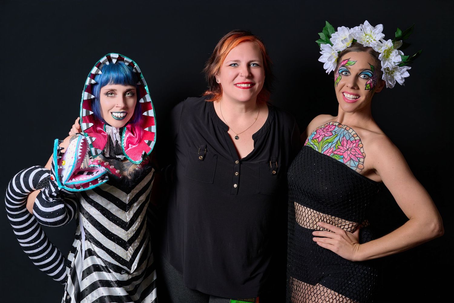 Three women pose together, two in colorful costumes with face paint and floral crown, one in casual black attire.