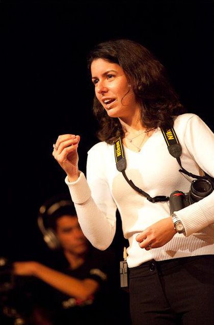 Woman in white sweater with Nikon camera strap gestures while speaking at an indoor event.