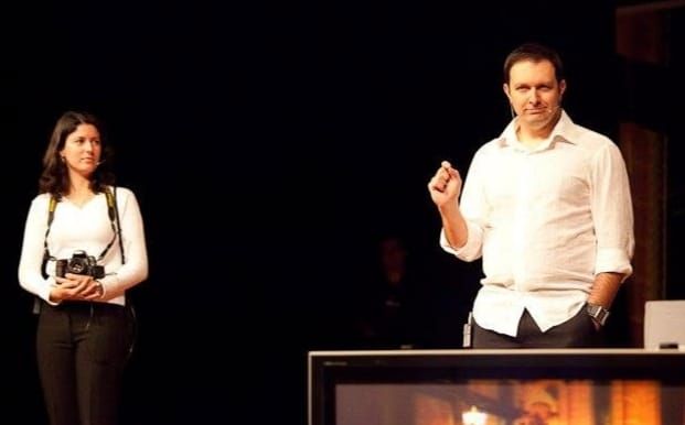 Man in white shirt gestures on stage while woman with camera stands nearby in dark theater setting.