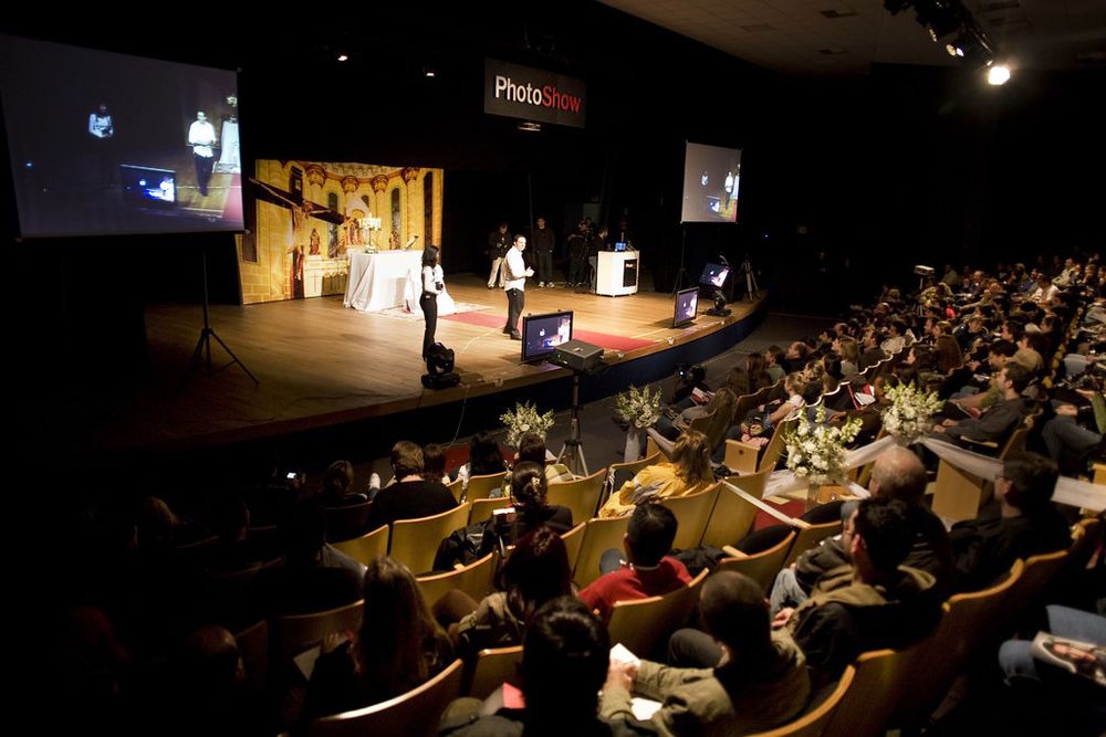 Large auditorium filled with audience watching a presenter on stage at a PhotoShop conference event.