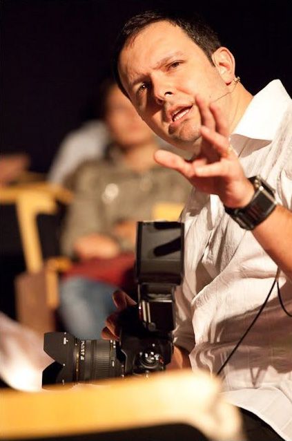 Man in white shirt holding a DSLR camera with flash, gesturing while speaking at a photography event.