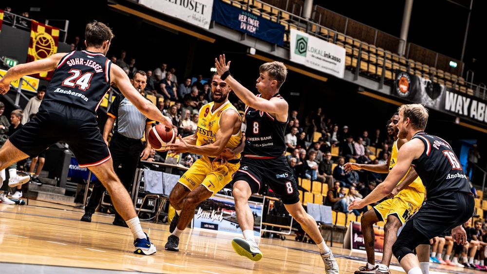 Basketball players compete in an intense game, with a yellow-jersied player driving through defenders on a bright indoor court.
