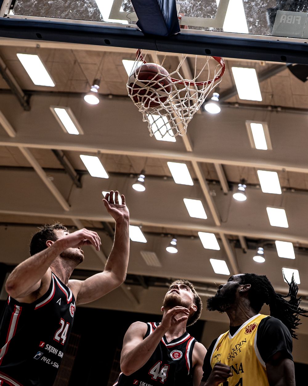 Basketball players in black and yellow uniforms compete for a rebound under the net during an indoor game.