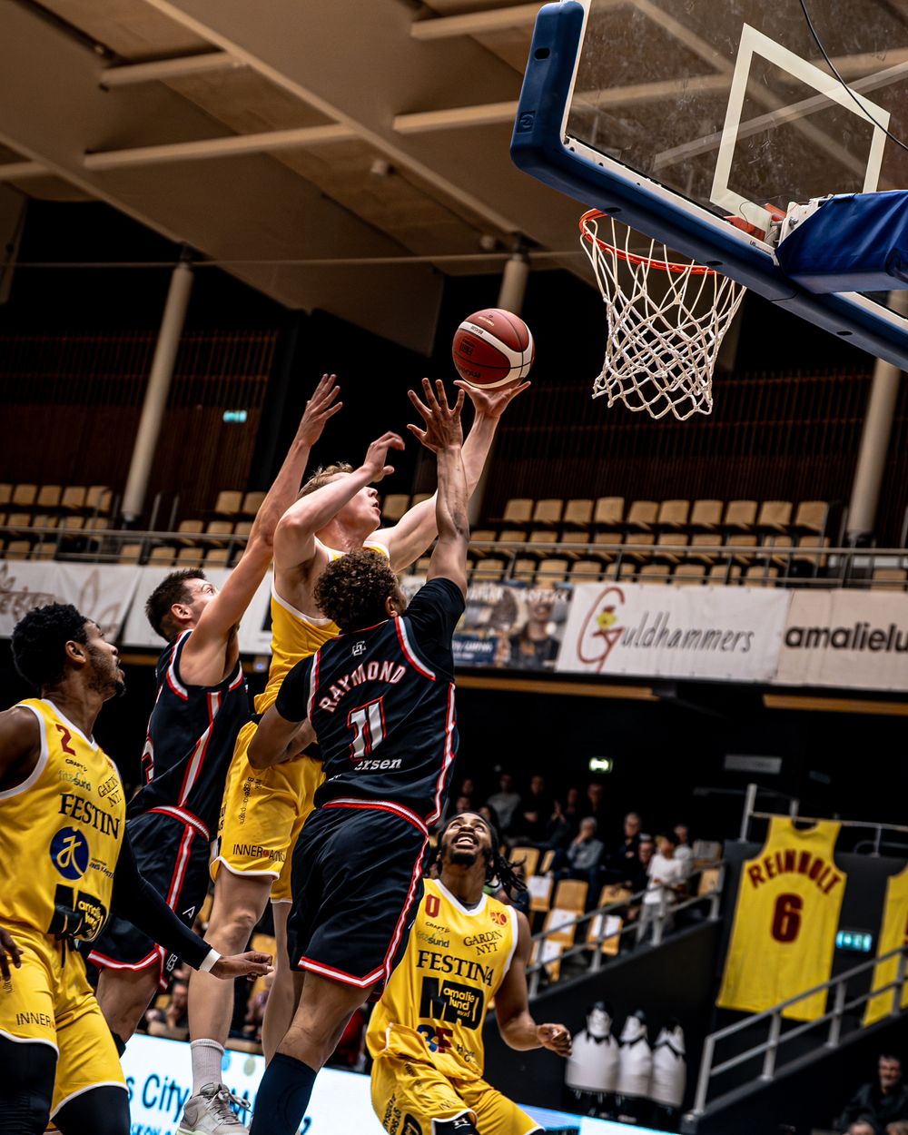 Basketball players in black and yellow uniforms compete for a rebound near the hoop during an indoor league game.
