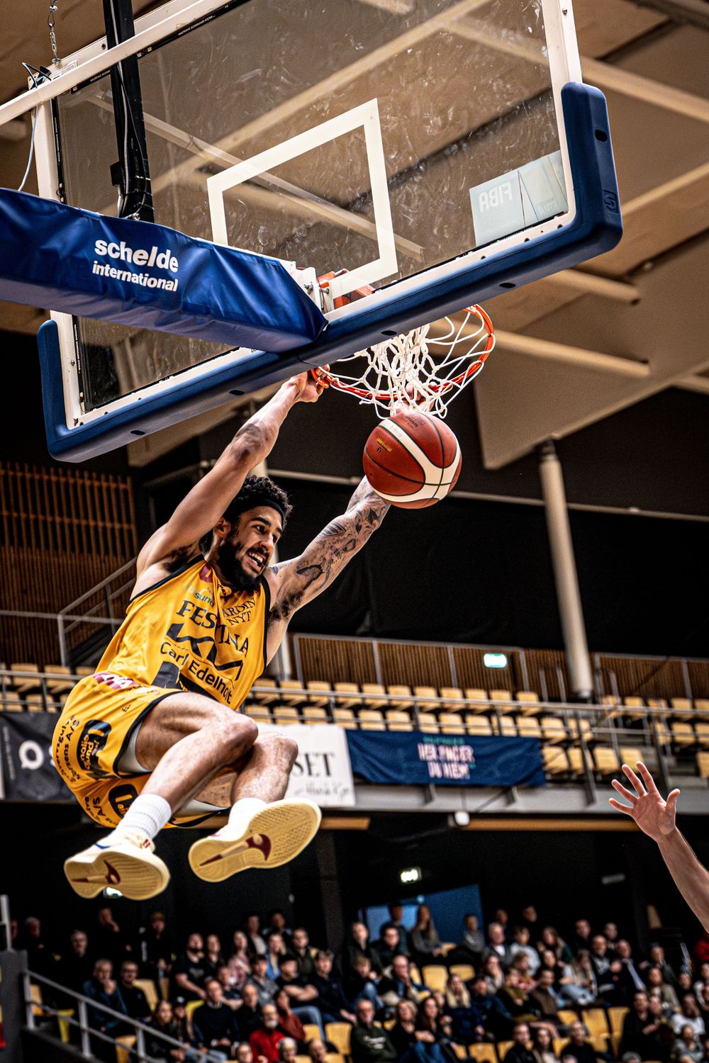 Basketball player in yellow jersey executes powerful slam dunk, hanging from rim during indoor game.